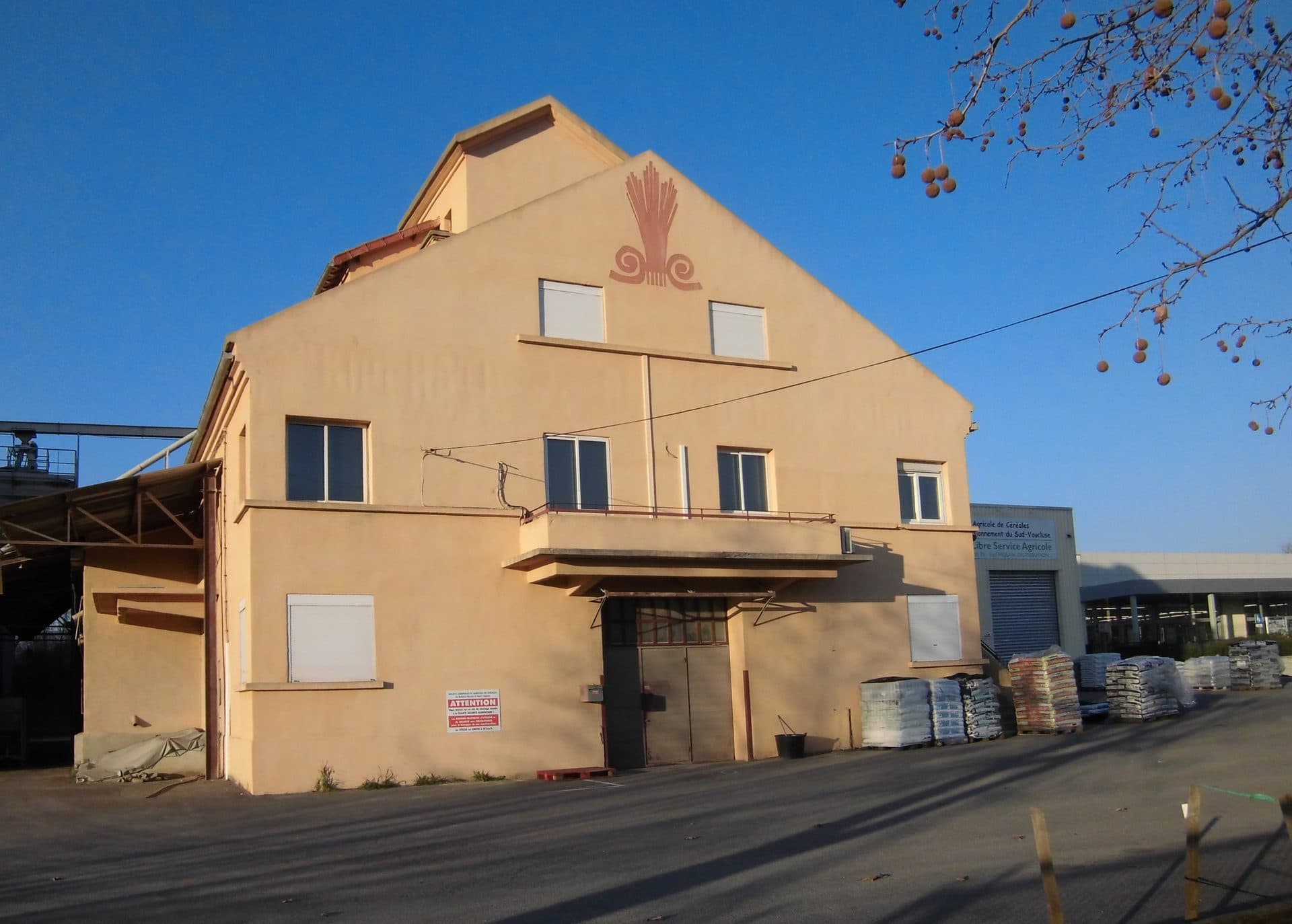 The Société Coopérative Agricole de Céréales grain cooperative at Apt in the Vaucluse, an ochre-rendered warehouse with an Art Deco wheat-sheaf motif on the gable, pallets of animal-feed and fertiliser sacks stacked on the forecourt beside its 'Libre Service Agricole' farm store