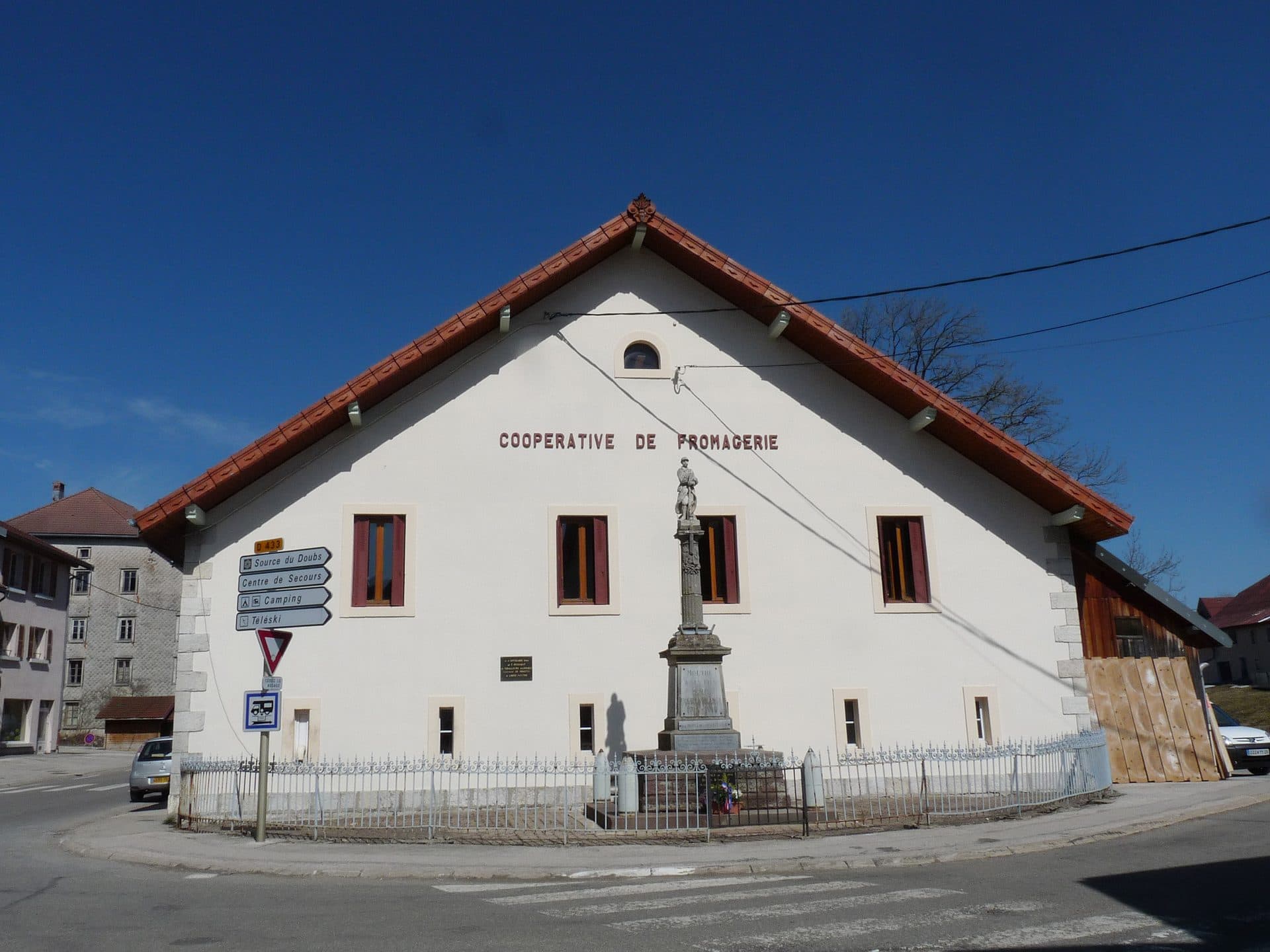 The village cheese cooperative at Mouthe in the Doubs — a white farmhouse-style building with a red tiled roof, 'COOPERATIVE DE FROMAGERIE' spelled across its gable in red lettering, with the commune's stone war memorial standing on the corner in front of it