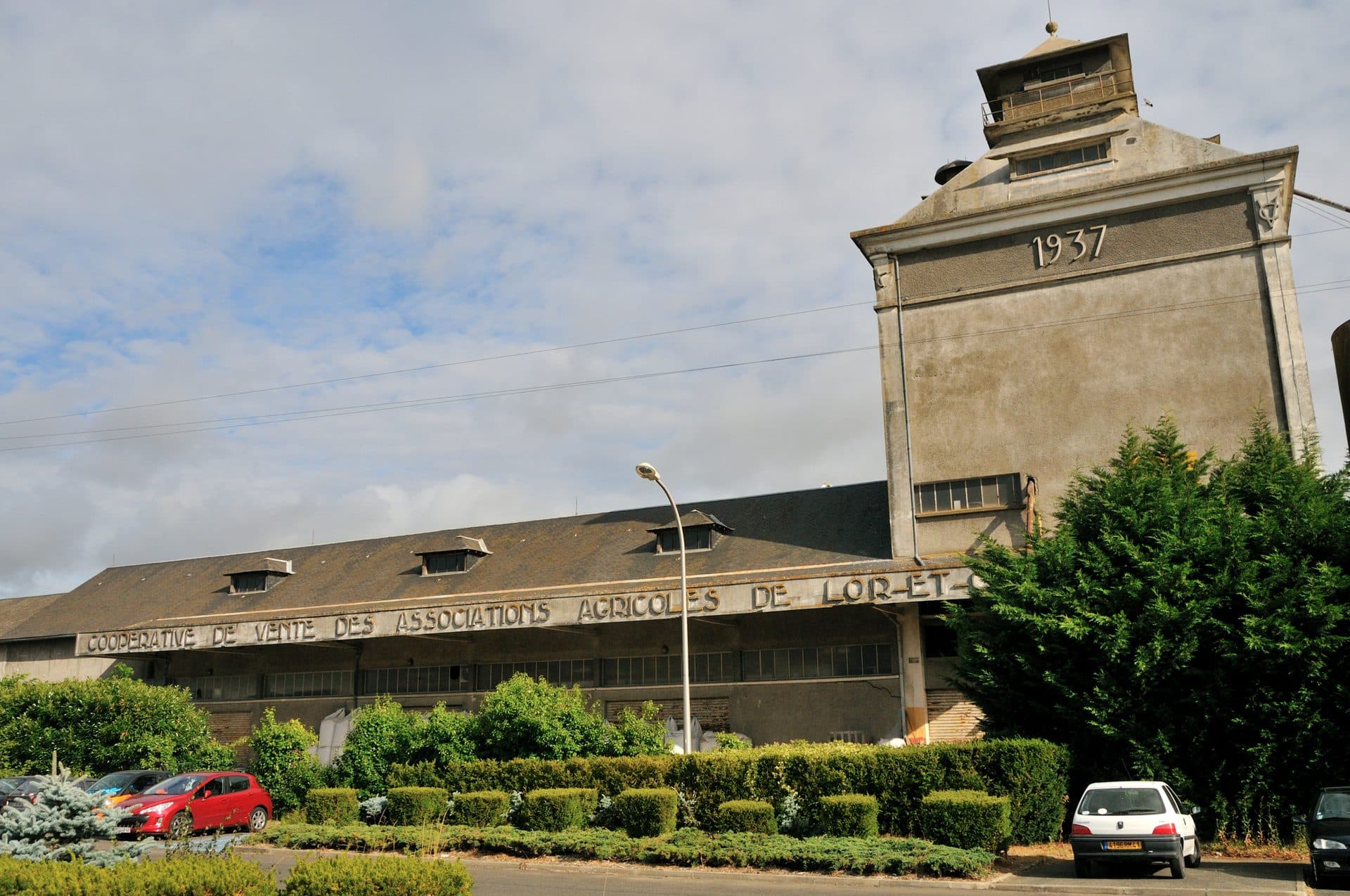 The historic 1937 grain silo of the Coopérative de Vente des Associations Agricoles de Loir-et-Cher at Onzain, its long warehouse facade carrying the cooperative's name in raised concrete relief lettering beneath a tall dated elevator tower