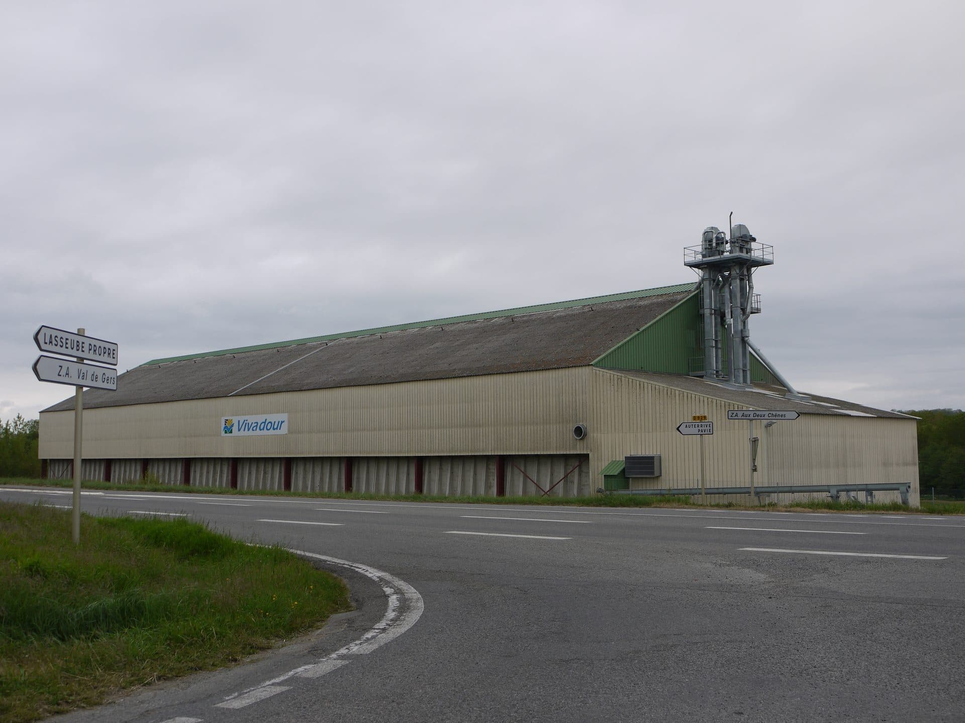 A long grain-storage warehouse of the Vivadour agricultural cooperative beside a road junction at Auterive in the Gers, its pale metal cladding carrying the Vivadour logo and a galvanised grain-elevator tower rising at one end