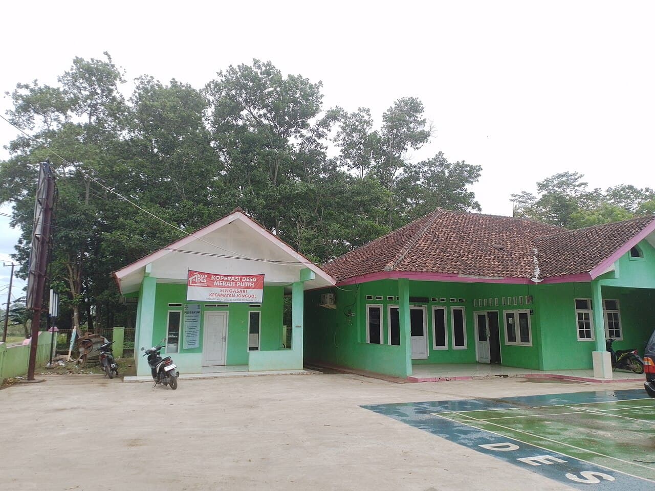 The village cooperative office of Koperasi Desa Merah Putih at Singasari in Jonggol district, West Java, Indonesia — a bright green single-storey building with a pink-trimmed gable and a banner across it naming the cooperative, motorbikes parked on the forecourt