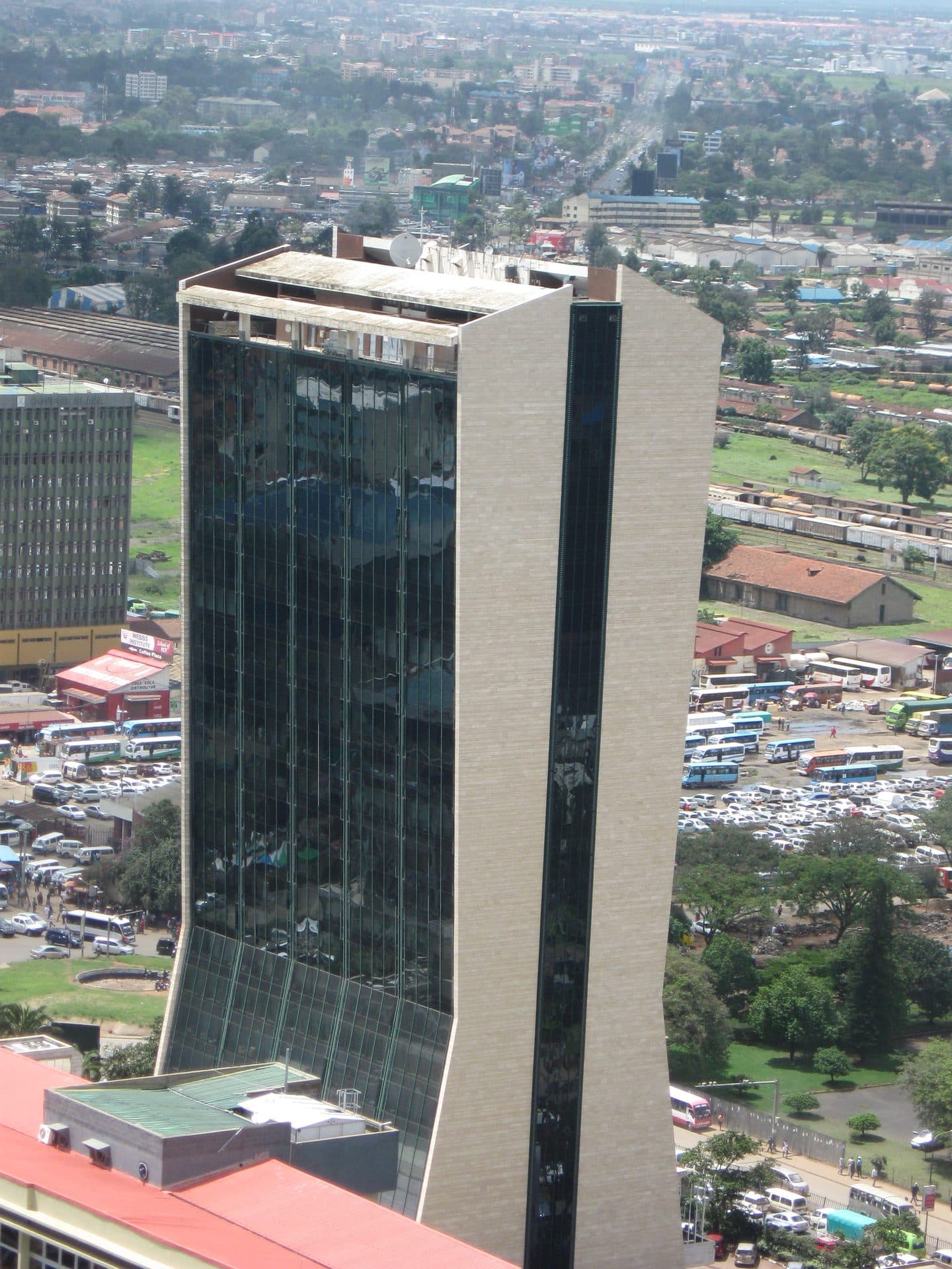 An aerial view of Co-operative House on Haile Selassie Avenue, Nairobi — the headquarters tower of the Co-operative Bank of Kenya, the bank majority-owned by Kenya's cooperative movement — its dark glass curtain wall and pale stone flank rising above the city's railway yards, matatu stage and rooftops