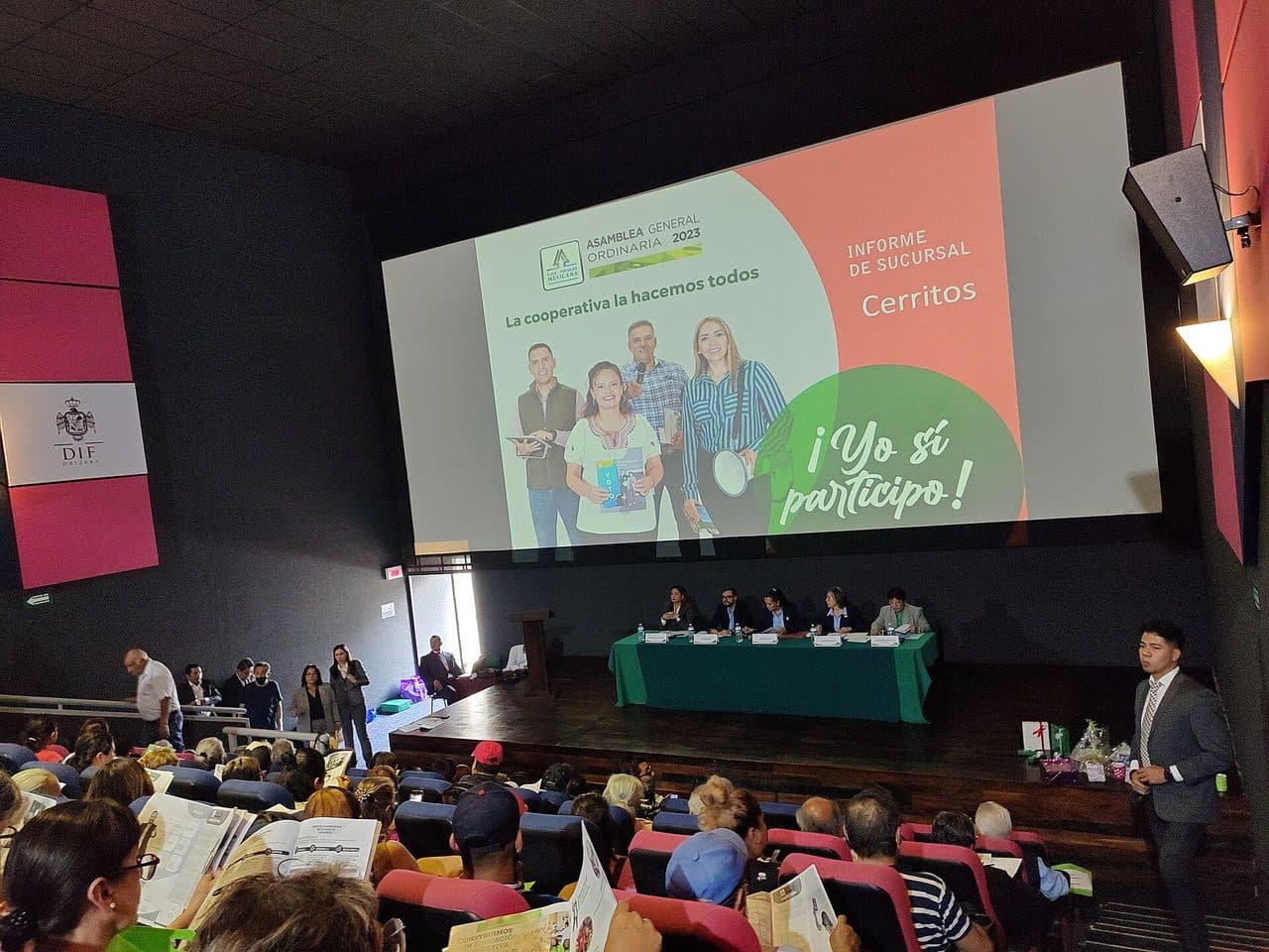 Members of Caja Popular Mexicana, Mexico's cooperative savings bank, at the 2023 ordinary general assembly of the Cerritos branch in Orizaba, Veracruz — rows of members holding printed branch reports in a cinema hall while the board sits at a green-clothed table beneath a screen reading 'La cooperativa la hacemos todos'