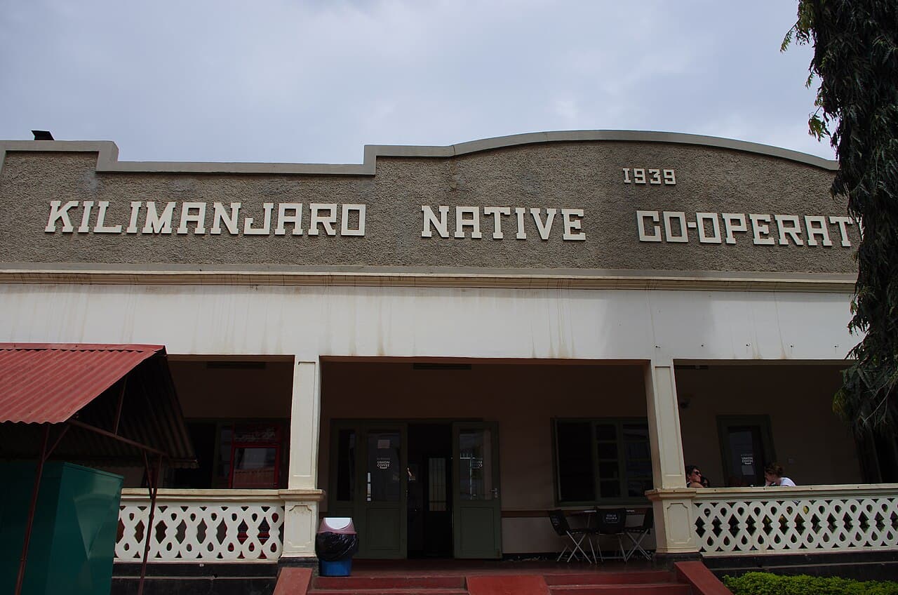 The 1939 headquarters of the Kilimanjaro Native Co-operative Union at Moshi, Tanzania — a pale Art Deco building with the coffee cooperative union's name and the date moulded in relief across its curved parapet, above an arcaded veranda now housing the Union Coffee café