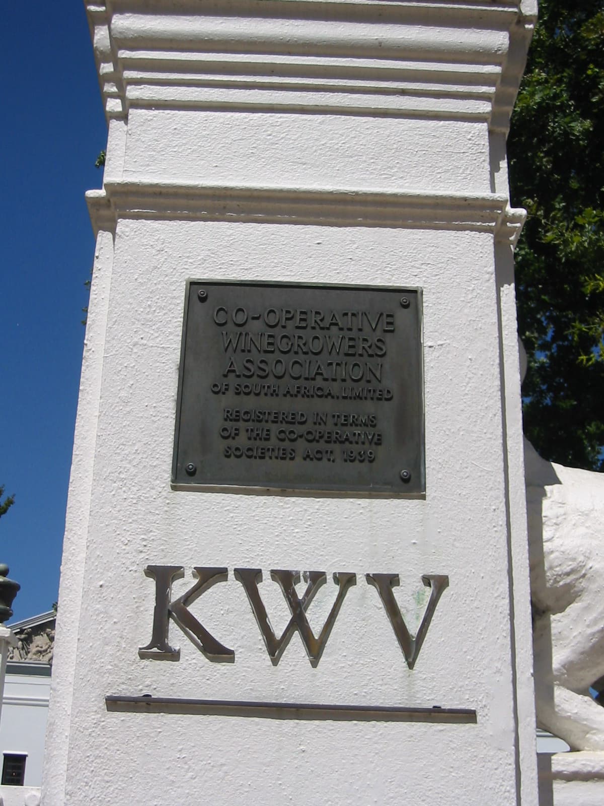 A bronze plaque on the whitewashed gatepost at KWV in Paarl, South Africa, reading 'Co-operative Winegrowers Association of South Africa, Limited — registered in terms of the Co-operative Societies Act, 1939', with the letters KWV mounted in metal below it against a deep blue sky