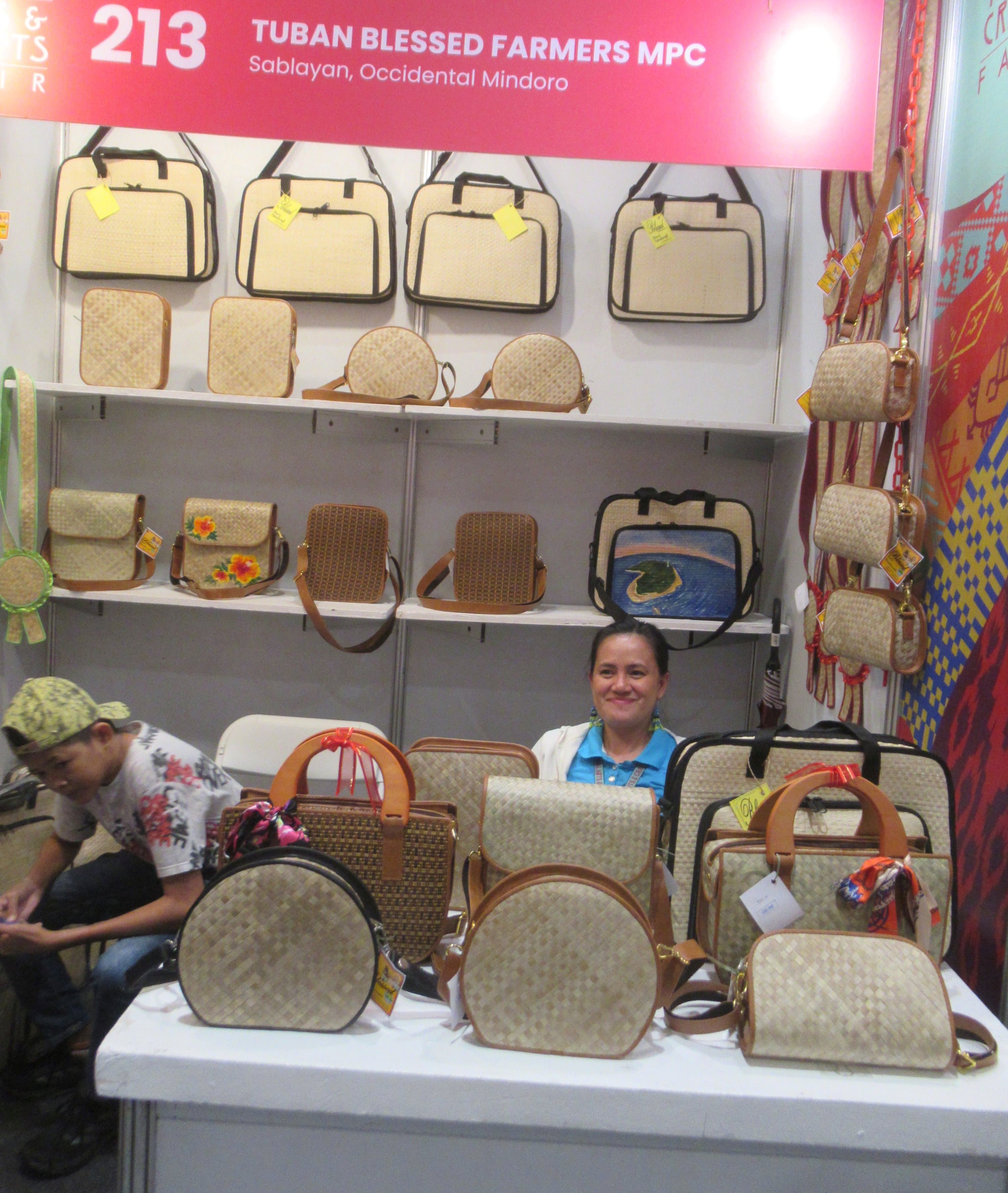 A booth for the Tuban Blessed Farmers Multi-Purpose Cooperative from Sablayan, Occidental Mindoro, at a Philippine crafts fair — a member sits behind a display of the cooperative's handwoven buri-palm handbags and purses